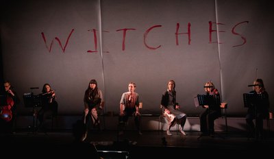 a group of women sit in a line on a stage in front of a large sheet of paper with Witches painted in red paint