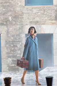 Sarah walks across a stage looking out over the audience, carrying 2 suitcases, wearing a head scarf