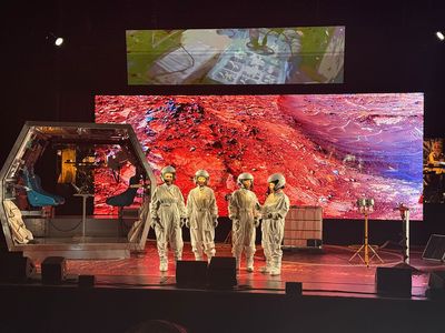 4 female cosmonauts stand in front of a metal space ship. 2 large rectangular screens can be seen hanging behind them, which are projecting images of space.
